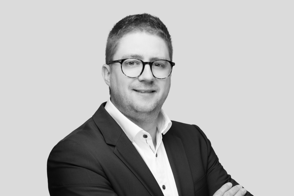 Porträt eines Mannes mit Brille und kurzem Haar in Anzug und Hemd, vor grauem Hintergrund.