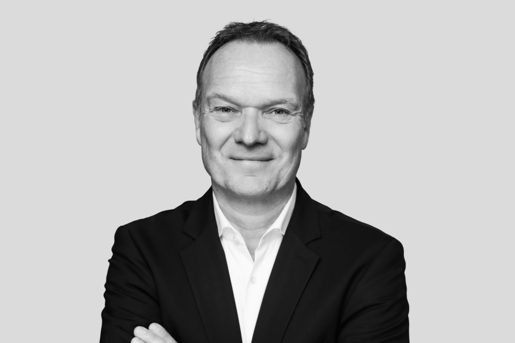 Porträt eines Mannes in dunklem Anzug mit verschränkten Armen vor grauem Hintergrund.