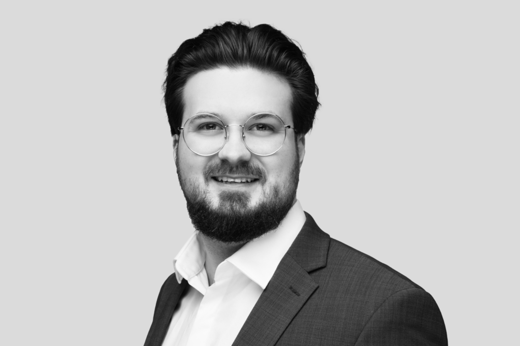 Porträt eines Mannes mit Vollbart, Brille und dunklem Anzug mit weißem Hemd.