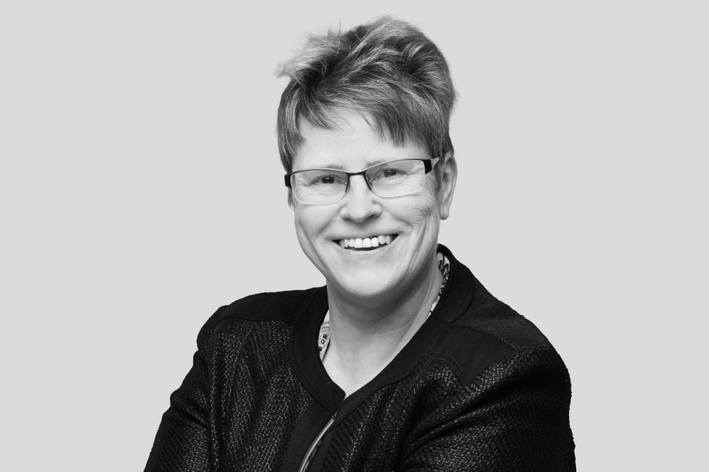 Porträt einer lächelnden Frau mit kurzer Frisur und Brille in dunkler Kleidung.