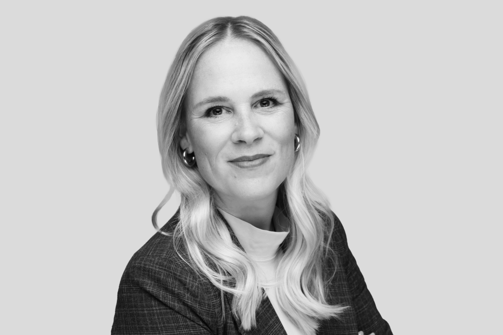 Porträt einer Frau mit langen, blonden Haaren in Businesskleidung vor grauem Hintergrund.