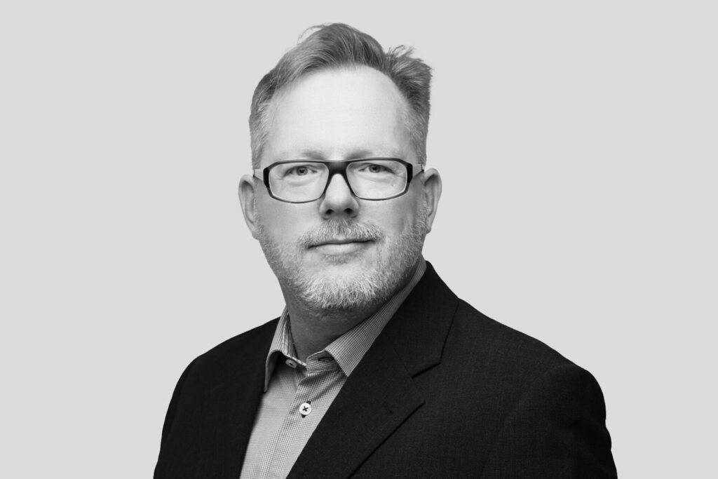 Porträt eines Mannes mit Brille, Bartansatz und Anzugjacke.